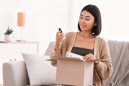 Happy asian lady unpacking parcel with beauty box Beauty Box Concept. Portrait of smiling asian woman received delivery package, holding foundation base for makeup, unpacking cardboard parcel with cosmetics product, sitting on the couch at home
