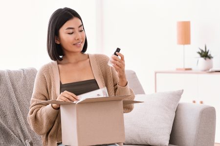 Happy asian lady unpacking parcel with beauty box Beauty Box Concept. Portrait of smiling asian woman received delivery package, holding foundation base for makeup, unpacking cardboard parcel with cosmetics product, sitting on the couch at home